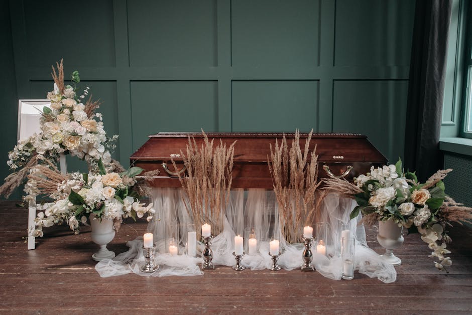 A floral arrangement for a funeral setting featuring two large white vases filled with cream and white roses, peonies, and greenery, complemented by pampas grass and dried flowers. The flowers are arr