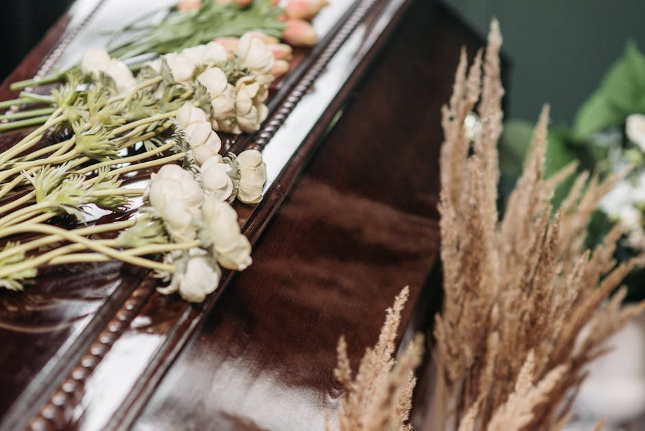 A close-up view of a floral arrangement placed on a polished wooden surface, featuring white ranunculus flowers with delicate, tightly packed petals and green stems, alongside tall, dried beige pampas