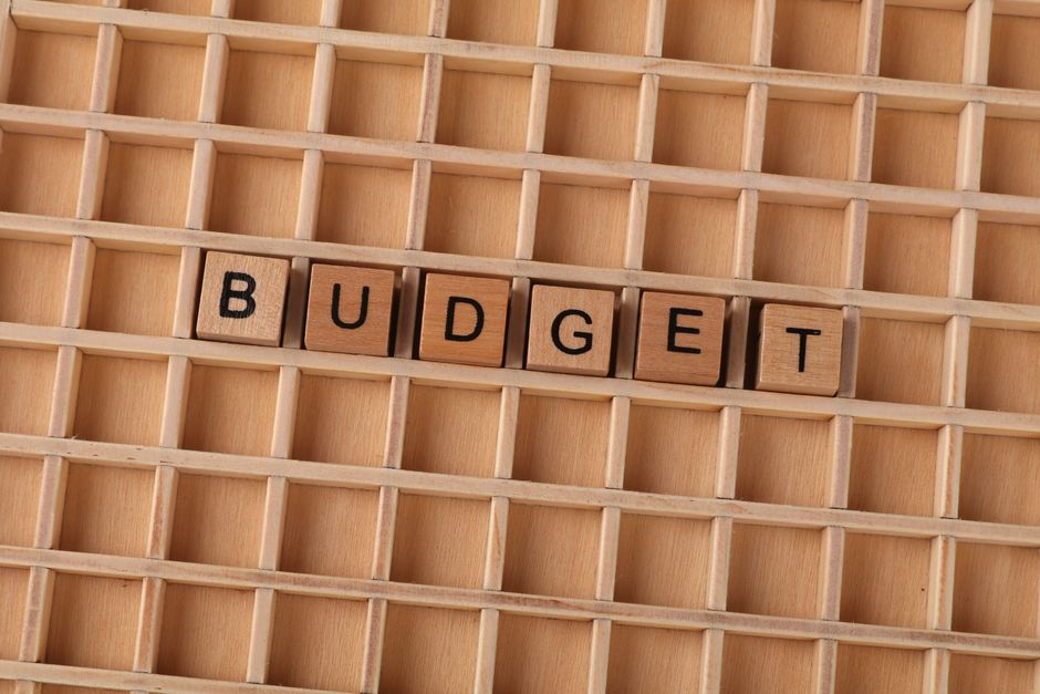 A wooden grid displaying small square compartments arranged in horizontal and vertical lines, with five wooden letter tiles spelling out 'BUDGET' placed in the middle row. The tiles are light brown wi