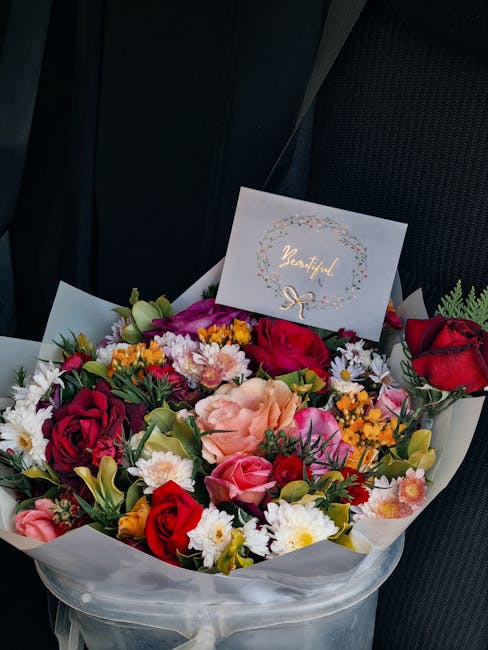 A vibrant bouquet of fresh flowers arranged in a circular style, featuring red roses, soft pink carnations, white daisies, yellow and purple blooms, and green foliage. The bouquet is wrapped in white