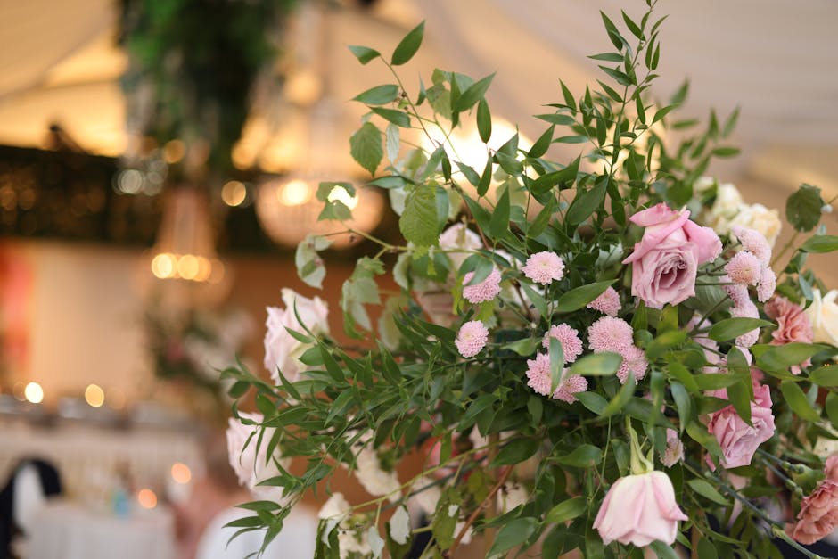 A floral arrangement featuring a mixture of soft pink roses, small pink chrysanthemums, and white flowers, all nestled among lush green foliage with slender, pointed leaves. The bouquet is arranged in