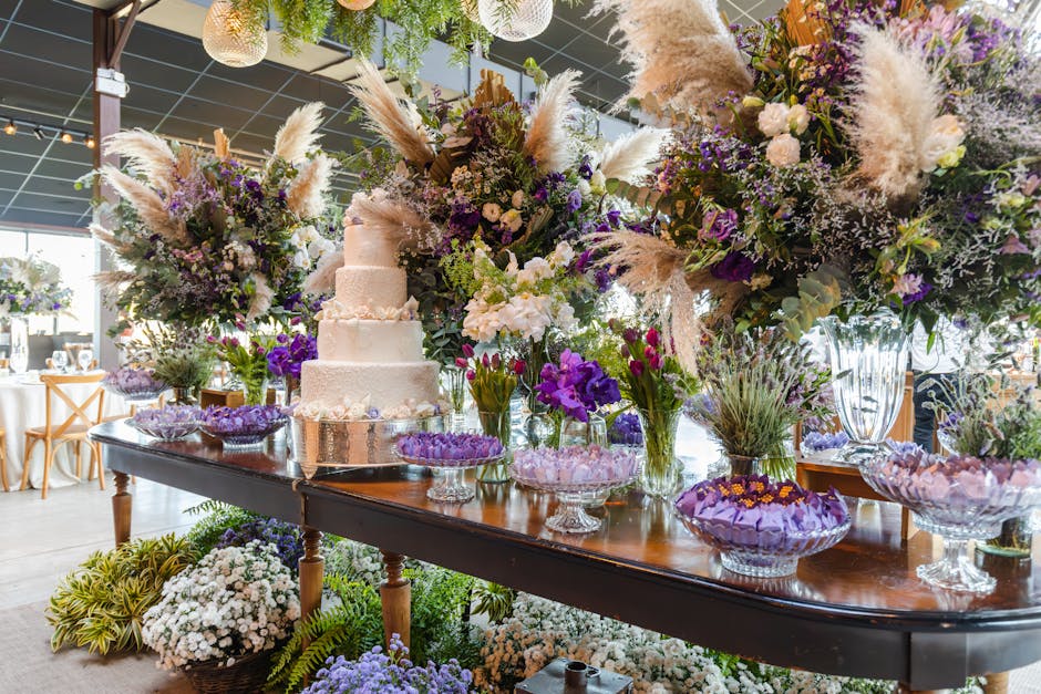 A floral arrangement display featuring large, lush bouquets of mixed flowers including white, purple, and lavender blooms with tall, feathery pampas grass accents. The flowers are arranged in clear gl