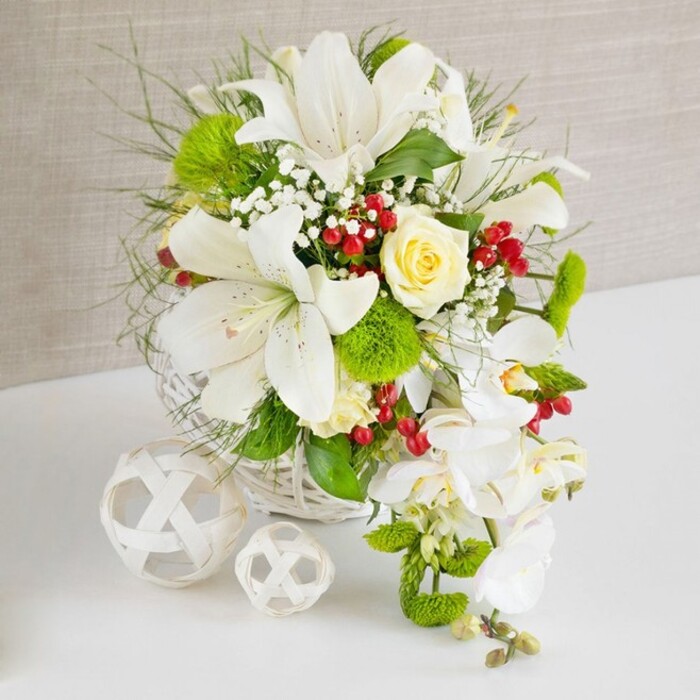An opulent arrangement of predominantly white blooms with lively green and red accents is displayed as a sophisticated table piece, reminiscent of a wedding or anniversary celebration in Southgate's quieter residential streets. The design is anchored in a round, white woven basket, its smooth wickerwork lending a modern yet organic foundation. Rising from the centre, several large Oriental lilies open generously, each bloom revealing gently recurved white petals dusted with fine speckles and surrounding a cluster of golden stamens. Just above them, a single creamy ivory rose, perfectly proportioned and delicately budded, provides a soft focal glow that contrasts beautifully with the crisp white lilies. Along the right side of the arrangement, white phalaenopsis orchids spill downward in a cascading line, their flat, elegant petals giving the piece a waterfall or teardrop appearance suited to a bridal table display near Southgate tube or a refined celebration at a local restaurant. Throughout the bouquet, fluffy green dianthus or pom-pom-style chrysanthemums nestle among glossy leaves and feathery foliage, adding depth, texture, and a fresh, grassy vibrancy. Ruby-red berries, likely hypericum, are dotted between the blooms, their polished surfaces catching the light and providing a festive accent. Tiny sprays of white gypsophila weave through the design like a mist, softening edges and filling gaps so that the arrangement appears full yet light. The entire display rests on a white tabletop whose faint reflection enhances the impression of cleanliness and care. To the left, two decorative white wicker balls, one larger and one smaller, echo the basket's weave, completing a serene scene that a local Southgate florist might prepare for same-day delivery to a special celebration at home.